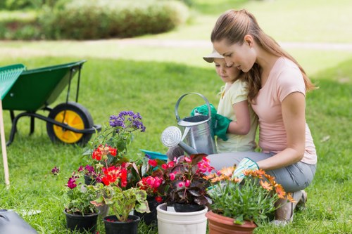 A woman and a young girl are crouched on a well-maintained grassy garden lawn in a suburban outdoor space, engaging in planting or tending to a variety of vibrant flowering plants and foliage arranged in pots. The garden features a lush, green, evenly cut lawn, with flower pots containing red, pink, purple, and orange blossoms, creating a colorful display. In the background, there is a hedge and a paved pathway, while a green garden wheelbarrow with yellow wheels is positioned to the left of the scene. The weather appears bright and sunny, casting natural light over the scene, and the overall environment is neat and inviting. This setting exemplifies routine outdoor care, suitable for professional gardening services such as those offered by Garden Maintenance Newham, focused on maintaining attractive and healthy gardens in the local area of Newham, UK.