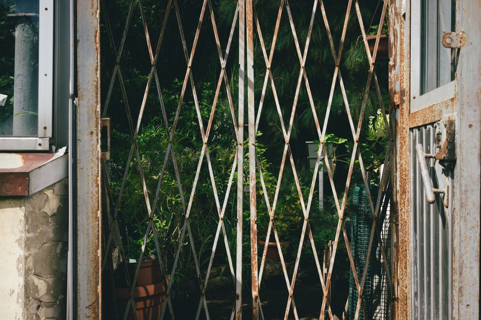 A rusty, metal garden gate with a diamond-shaped lattice pattern, partially open to reveal a lush garden beyond. The garden contains dense green foliage, including various plants and shrubs with leave