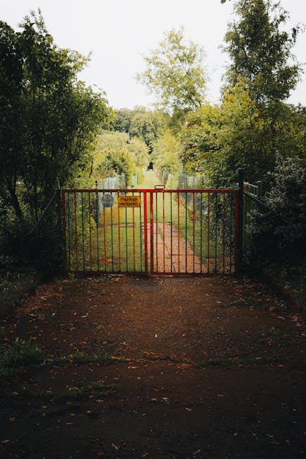 A rusty, metal garden gate with a diamond-shaped lattice pattern, partially open to reveal a lush garden beyond. The garden contains dense green foliage, including various plants and shrubs with leave